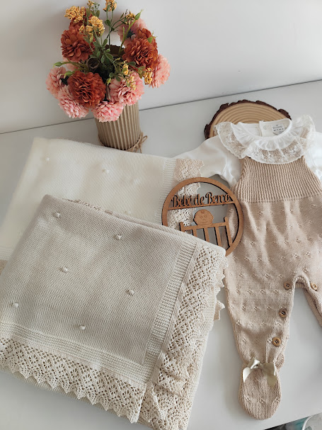 Manta cremosa com renda e roupa de bebé marrom claro com gola rendada branca, com flores artificiais ao lado