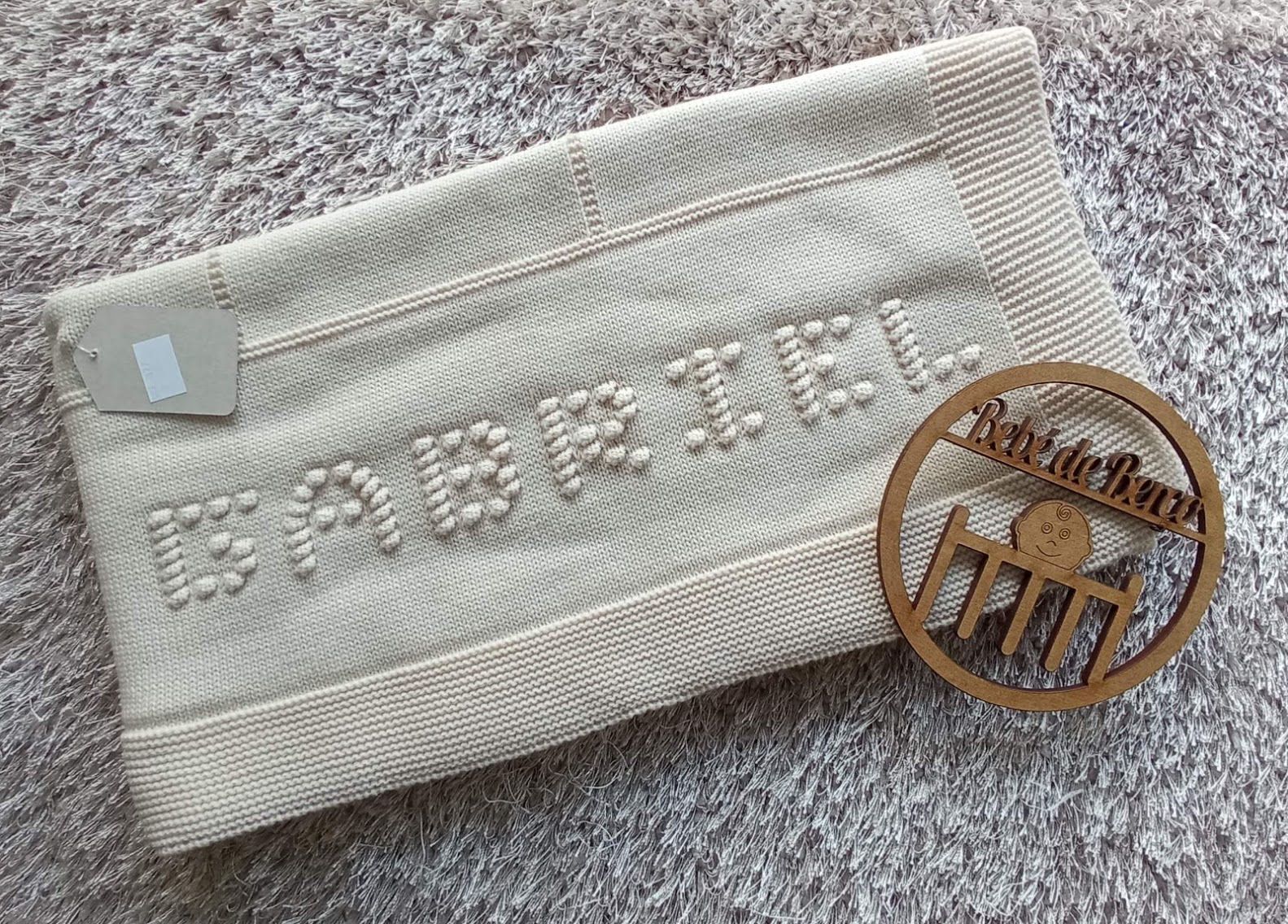 Manta bege clara com nome 'GABRIEL' em pérolas, sobre tapete cinza, com etiqueta e suporte de madeira