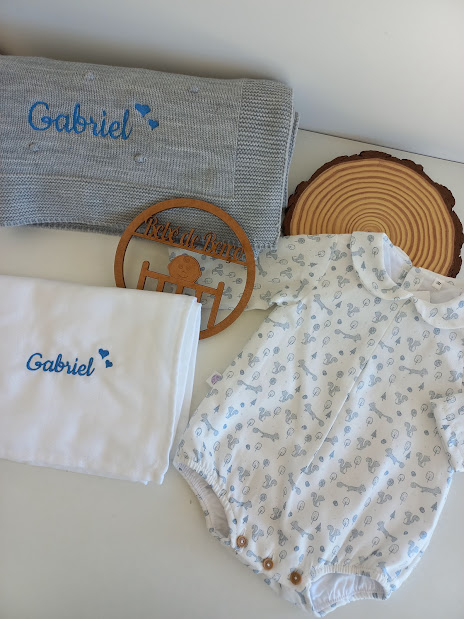 Conjunto de roupa para bebé com body, manta e pano branco com bordados azuis e objeto decorativo de madeira