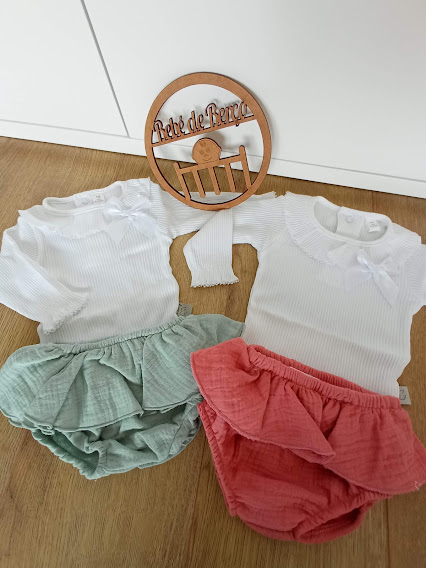 Dois conjuntos de roupa de bebé com saia verde e rosa e corpos brancos em chão de madeira