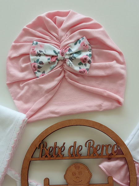 Touca turban rosa com laço floral e objeto de madeira com texto Bebé de Berço