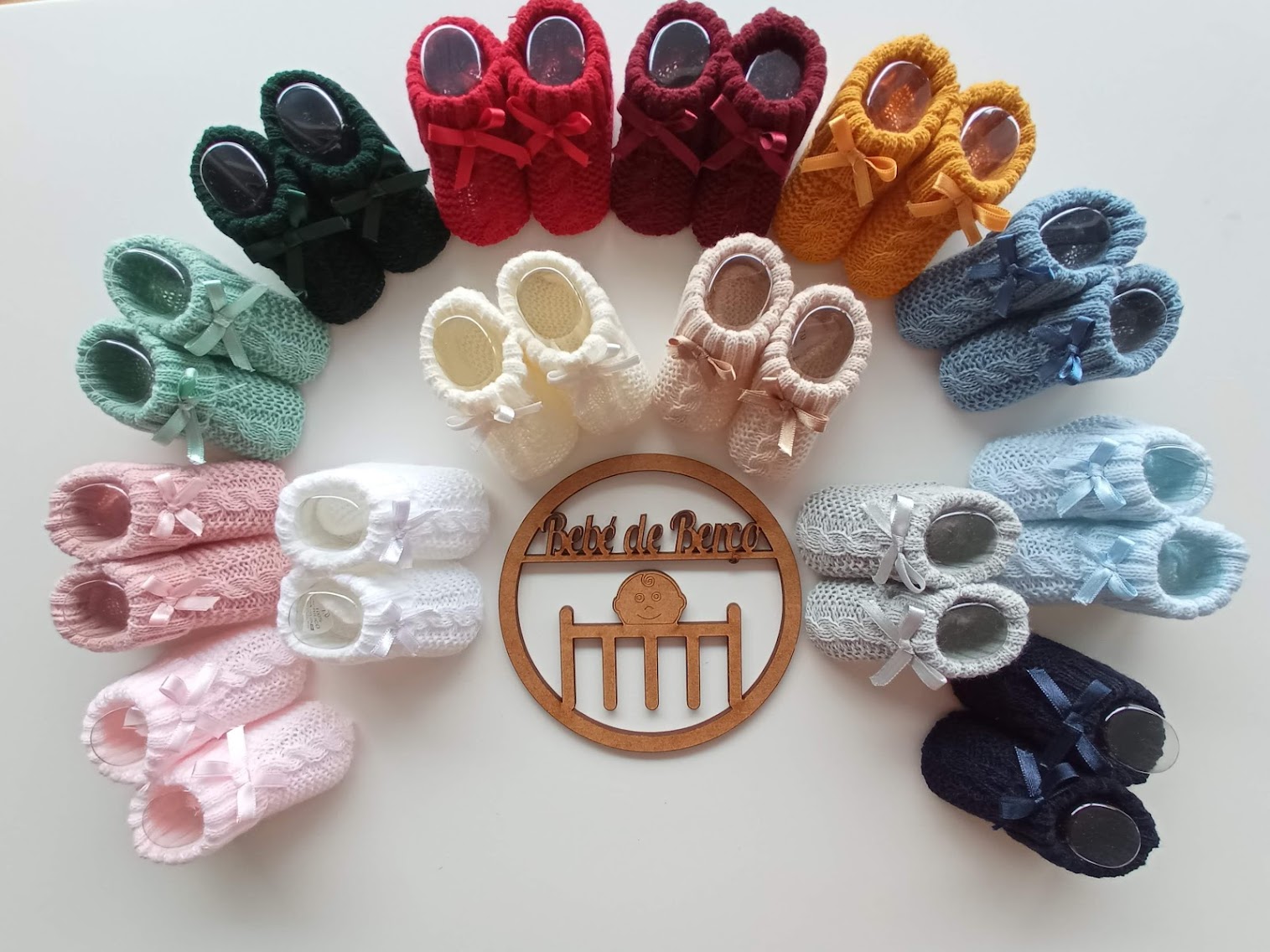 Sapatinhos de bebé de tricô em diversas cores dispostos em círculo com um símbolo de madeira no centro