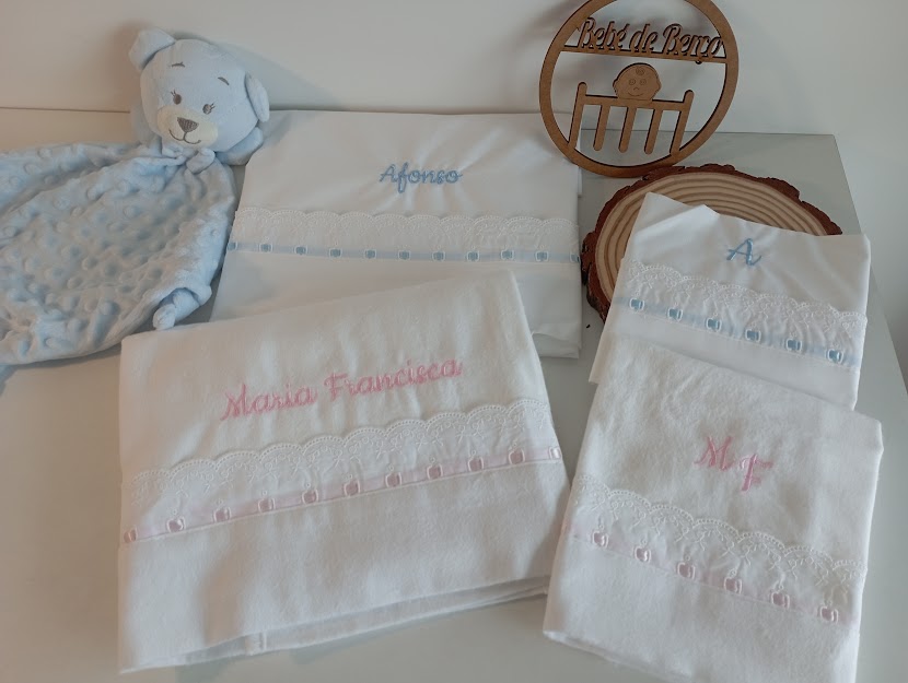Paninhos de bebê brancos bordados com nomes, manta azul macia e placa de madeira com texto Pé de Beira