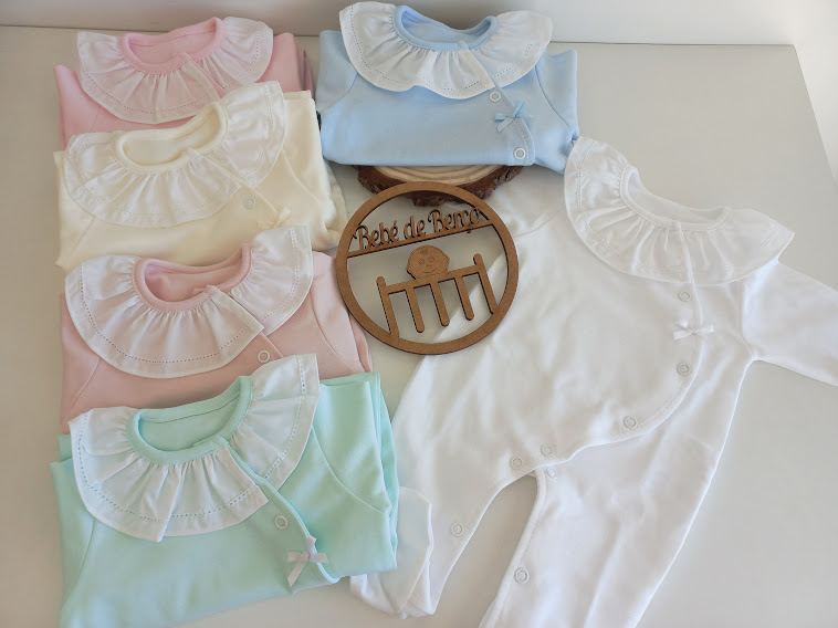 Bodies para bebé de várias cores com golas rendadas brancas e placa redonda de madeira com texto.