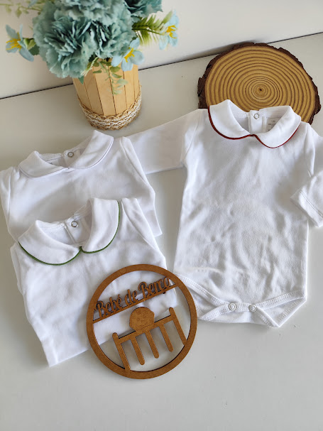 Três bodies brancos de bebé com golas bordadas e ornamento de madeira com texto junto a planta numa mesa branca