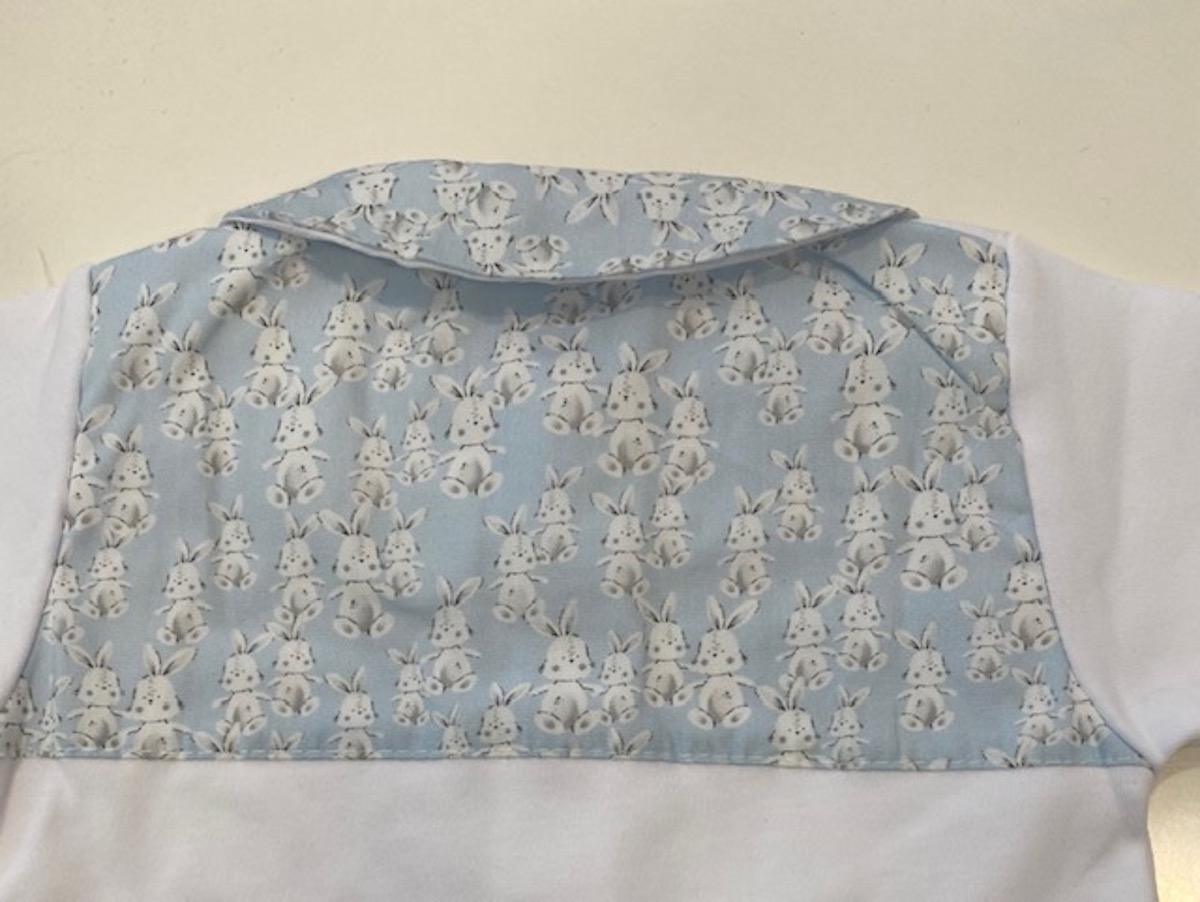 Peça de roupa infantil branca com tecido azul claro estampado com coelhos na parte superior e gola virada
