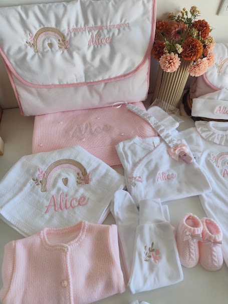 Conjunto de roupa de bebé branca e rosa com bordados e acessórios organizados numa mesa com flores