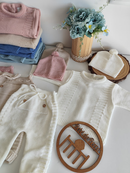Conjunto de roupa de bebé em malha com camisolas, calças e gorros coloridos, ao lado de um vaso de flores azuis.