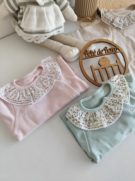 Três pijamas de bebé com golas rendadas em tecido estampado, em cores rosa, verde e bege, dispostos sobre uma superfície branca com boneca de peluche e placa de madeira Bebé de Berço.