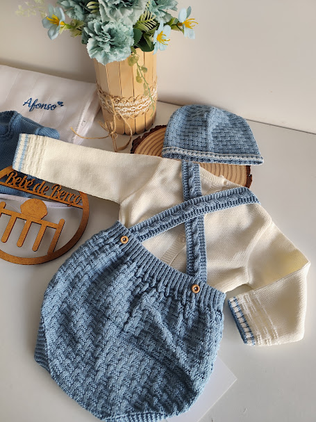 Conjunto de bebé tricotado creme e azul com camisola, calções com alças e gorro, em fundo branco com decoração