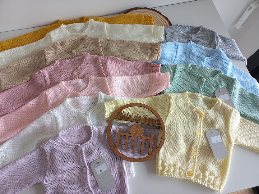 Oito casacos de malha de bebé em várias cores claras com botões e etiquetas.