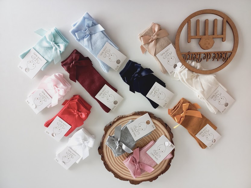 Vários pares de meias de bebé de cores sólidas com laços e etiquetas sobre uma mesa clara com objetos decorativos