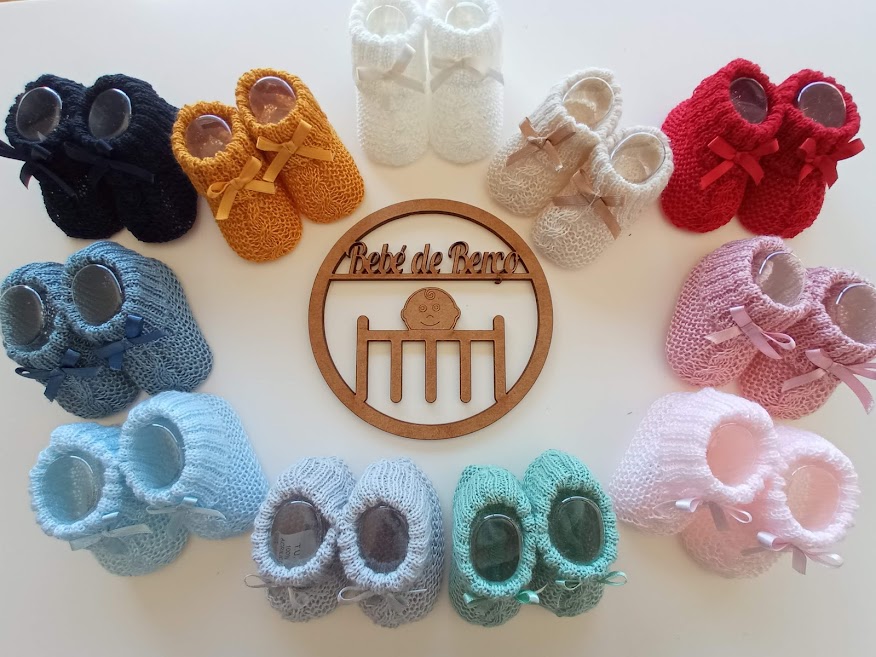 Sapatinhos de bebé de malha de várias cores dispostos em círculo sobre uma mesa branca com uma placa de madeira ao centro.