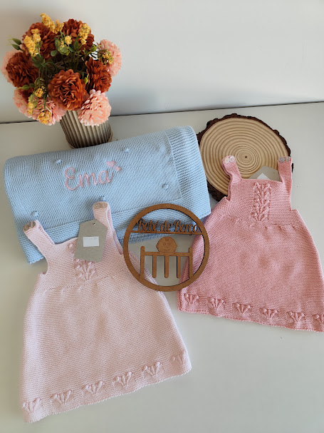 Dois vestidos de malha rosa claro, cobertor azul com nome bordado, vaso com flores secas e ornamento de madeira.