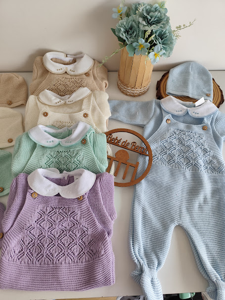 Conjunto colorido de roupa de bebé em malha com padrão trançado e gorros, em fundo claro com vaso de flores