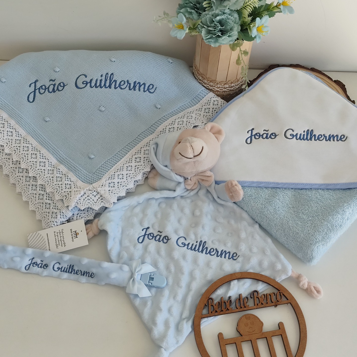 Conjunto de produtos de bebé azul e branco personalizados com o nome João Guilherme junto a uma jarra de flores
