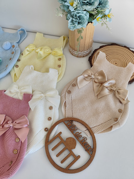 Várias peças de roupa para bebé em malha com botões e laços, dispostas junto a flores e ornamento de madeira