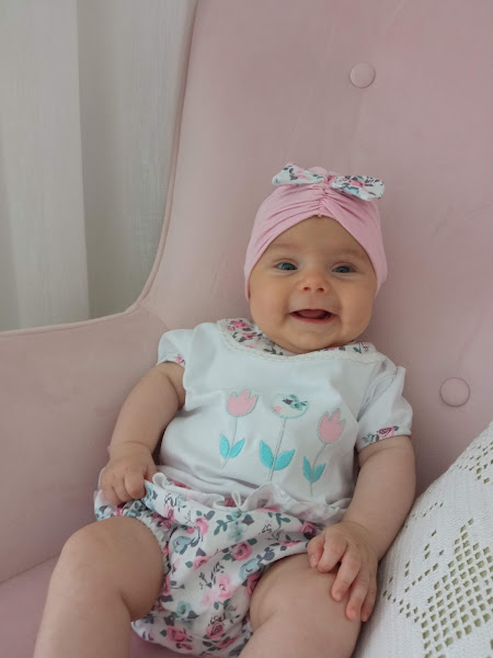 Bebé com roupa branca com flores, calções floridos e touca rosa sentada numa cadeirão cor de rosa.