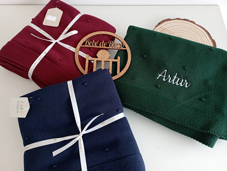 Três mantas de bebê coloridas e amarradas com fitas ao lado de placa de madeira com texto