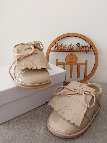 Sapatos de bebé bege claro em verniz com atacadores e franja, sobre caixa branca e placa madeira com texto