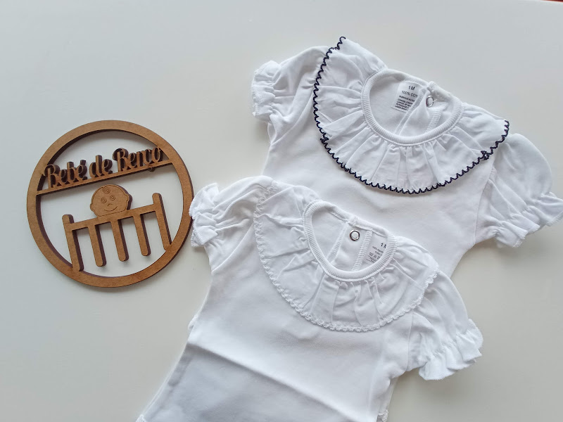 Dois bodies brancos para bebé com golas frisadas e placa de madeira com texto e imagem de berço