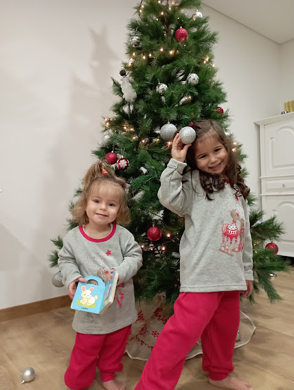 Duas crianças com pijamas vermelhos e cinzentos junto a uma árvore de Natal decorada com bolas e luzes.