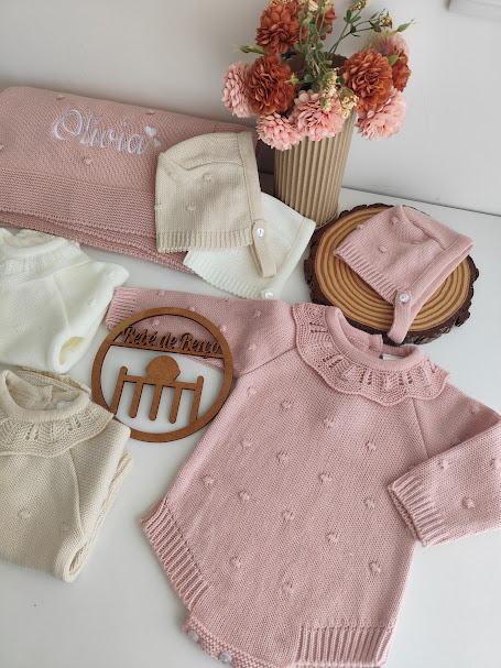 Roupas de bebé em malha rosa, creme e branco com detalhes e um arranjo de flores