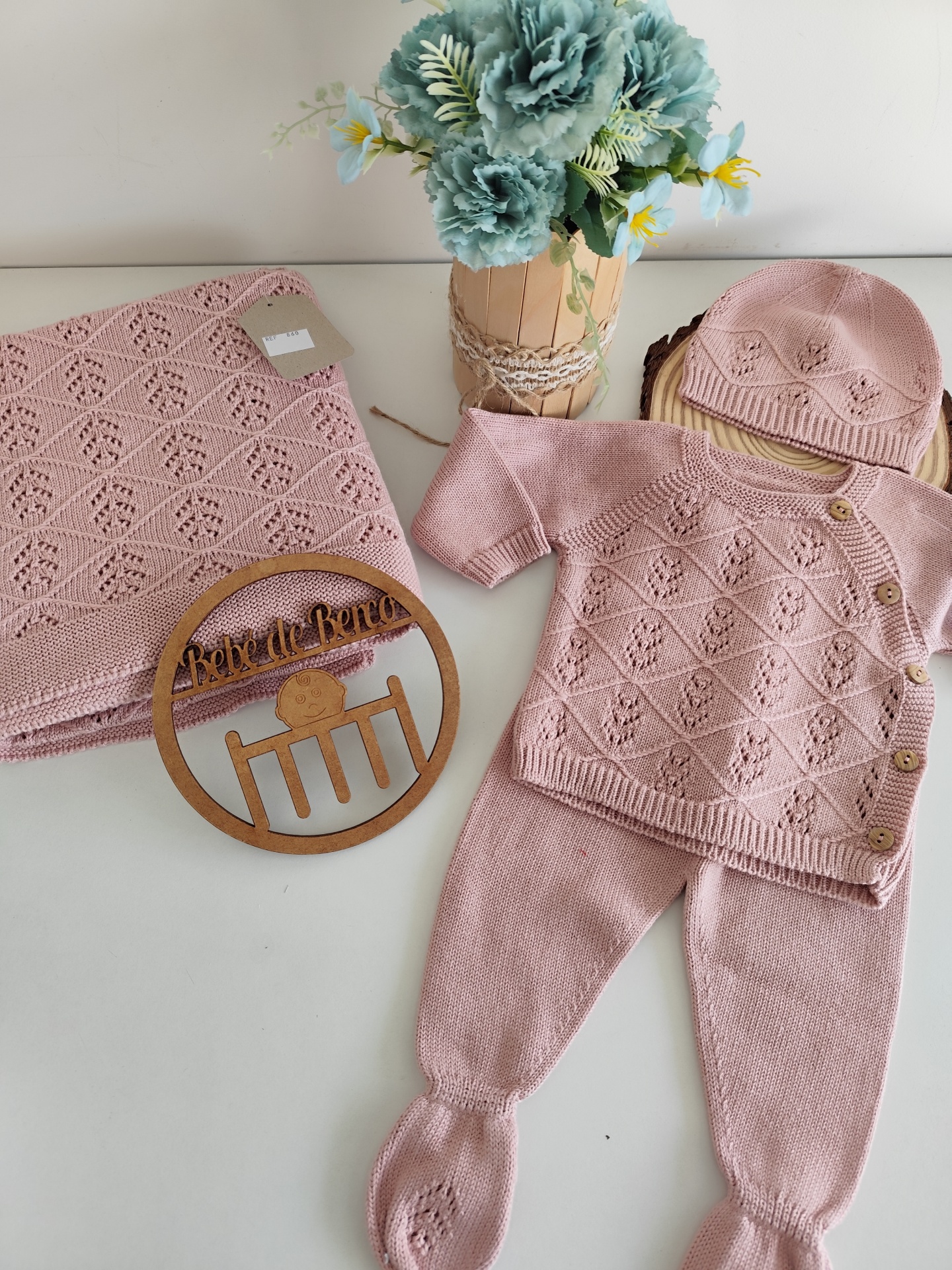 Conjunto de bebé de malha rosa com camisola, calças e gorro ao lado de manta e jarro de flores