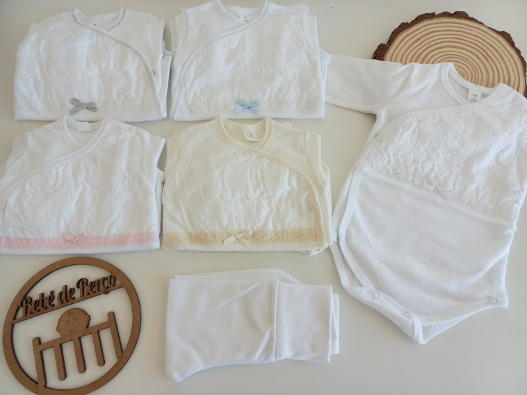 Conjunto branco de roupa de bebé com detalhes em renda e laços coloridos sobre superfície branca e suporte de madeira com texto