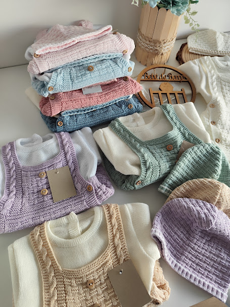 Roupas de bebê em malha em vários tons pastel com botões de madeira sobre mesa branca com vaso de flores e sinal de madeira