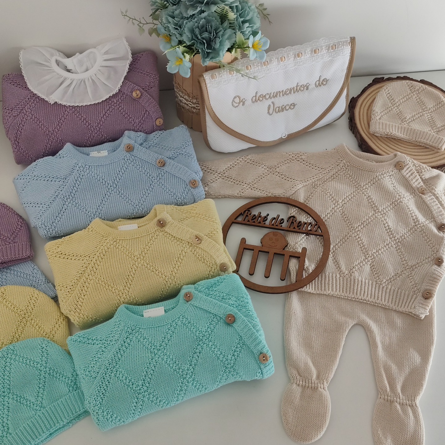 Conjuntos de roupa de bebé em tricô de várias cores pastéis com botões de madeira e um saco branco bordado
