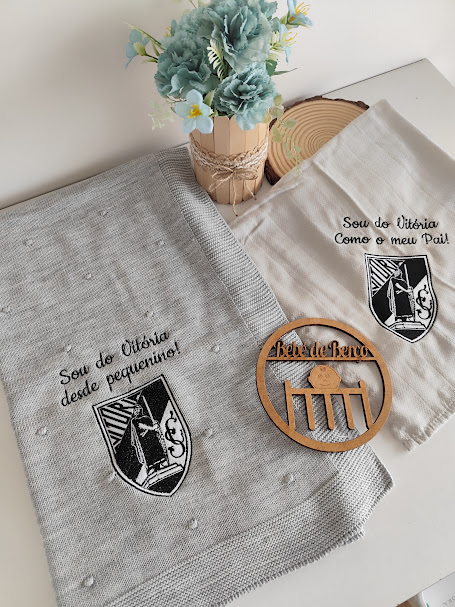 Tecidos bordados com brasão do Vitória, vaso de flores e emblema de madeira sobre superfície clara.