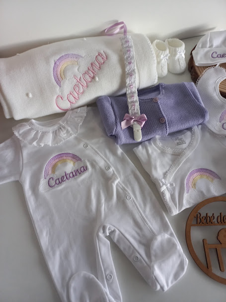 Conjunto de roupa de bebé branca e lilás com bordados de arco-íris e nome Caetana