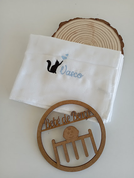 Toalha branca com bordado azul e preto e enfeite de madeira com texto 'Bebé de Berço'