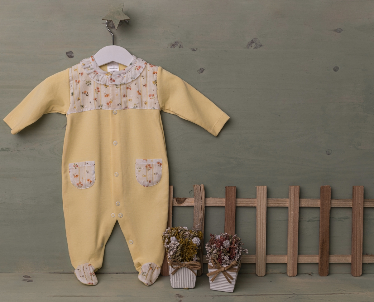 Babygrow amarelo com detalhes florais e botões, pendurado com cerca de madeira e vasos de flores secas ao lado