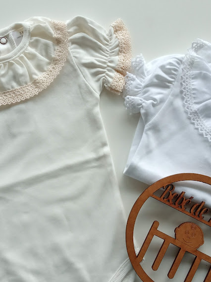 Dois bodies infantis, um creme com rendas bege e outro branco com rendas brancas, com círculo decorativo de madeira.