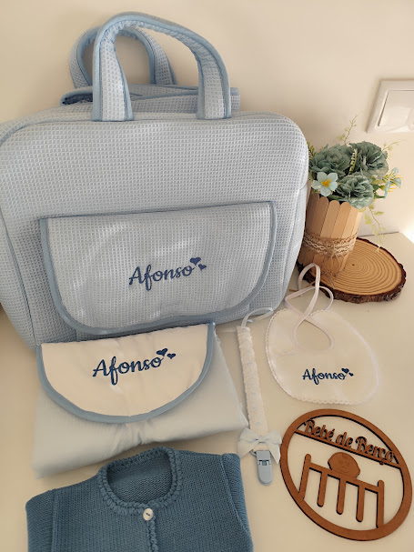 Conjunto de acessórios de bebé com mala azul claro, babete, cobertor, chupeta e camisola, com nome Afonso bordado, e placa em madeira com texto Retiró de Renê