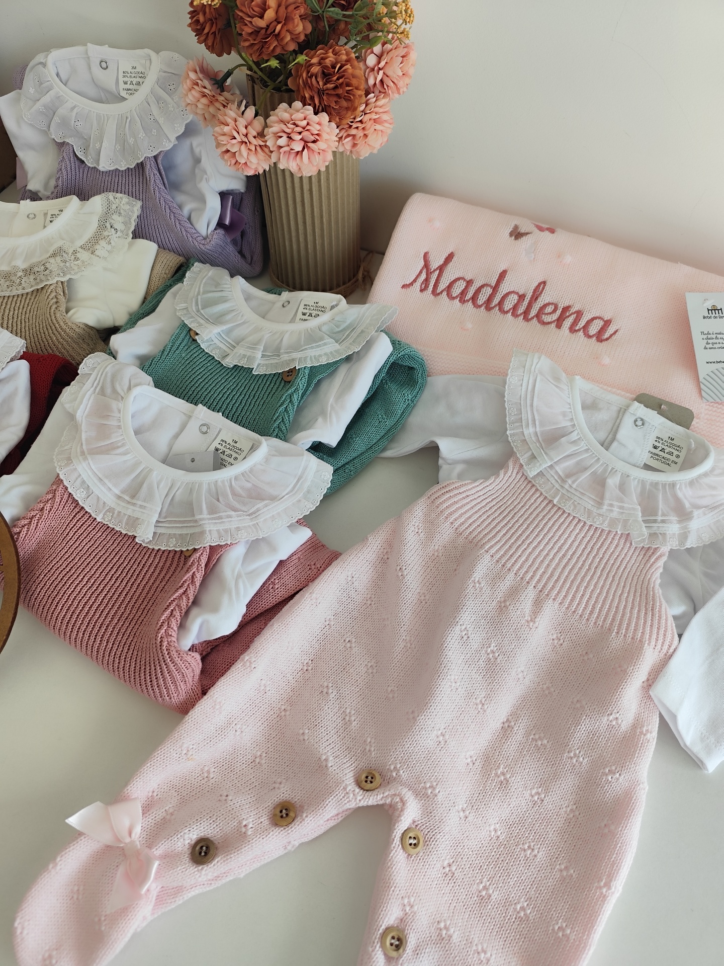 Roupas de bebé em malha e algodão coloridas com gola de renda branca, botões de madeira e cobertor rosa bordado com 