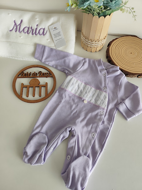 Macacão de bebé lilás com botões e faixa branca, toalha branca bordada com 'Maria' e decoração em madeira e flores