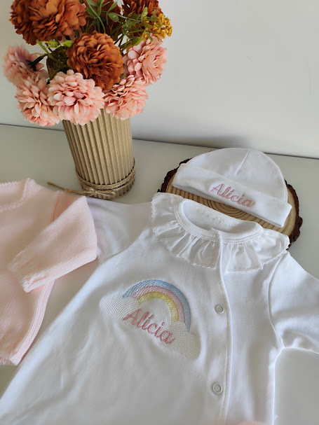 Roupinha de bebé branca com bordado 'Alicia' e flores artificiais num vaso