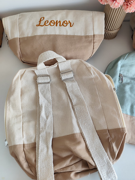Mochila e bolsa bege e castanho, bolsa com nome bordado Leonor, vaso com flores rosa no fundo