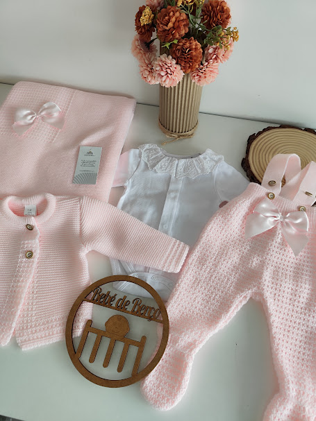 Conjunto de roupa de bebé rosa e branca com manta, corpo e calças, junto a vaso com flores secas
