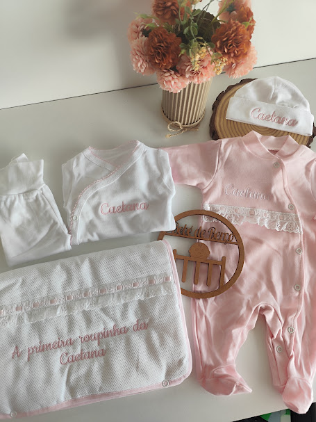 Conjunto de roupa de bebé rosa e branca com bordados, manta e gorro, sobre mesa branca com flores em vaso
