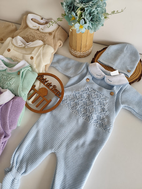 Conjunto de roupa de bebé em malha azul claro com gorro e vários conjuntos coloridos ao fundo num cenário branco com vaso de flores