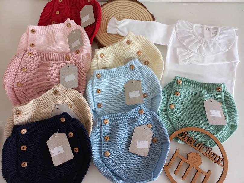 Conjunto colorido de roupas de bebé em malha e body branco com golinha, etiquetas penduradas, boneco de madeira Kids & Beans