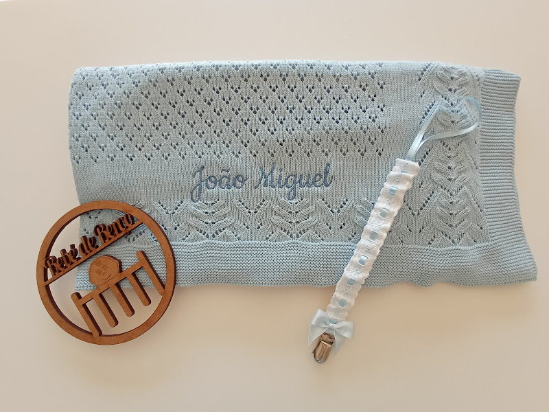 Manta azul clara com nome bordado e chupeta com fita e prendedor metálico, além de ornamento redondo de madeira com texto