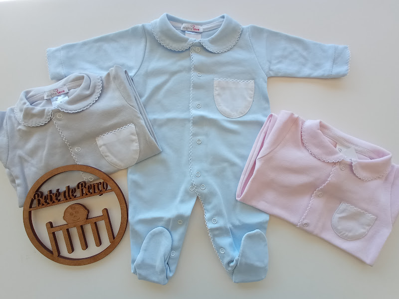 Três macacões de bebé nas cores azul, cinza e rosa com gola redonda e bolso frontal, junto a uma placa de madeira gravada.