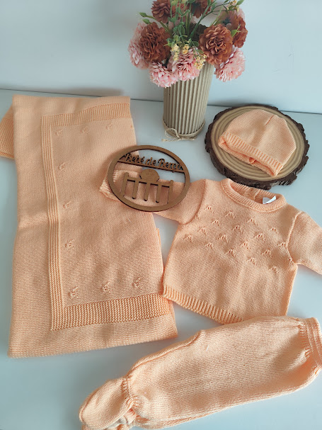Conjunto roupa bebé malha pêssego com manta, camisa, calças e gorro junto a flores e base de madeira