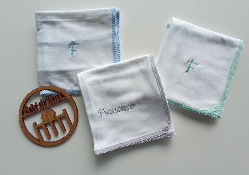 Três fraldas brancas com bordas coloridas e bordados, e uma placa redonda de madeira sobre fundo branco