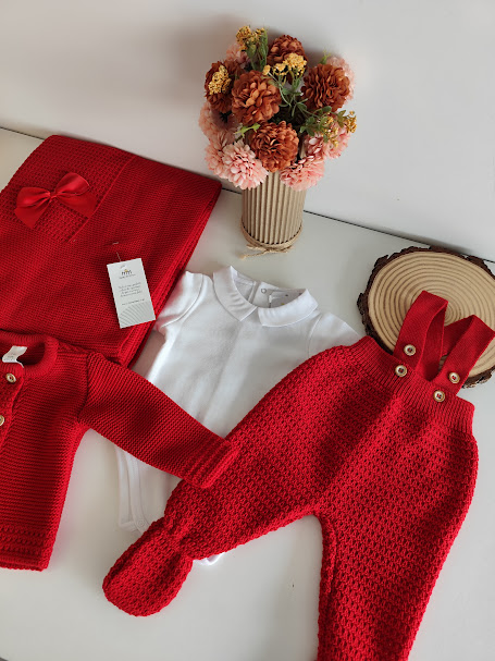 Conjunto de roupa de bebé vermelha e branca em malha, com manta, sobre mesa branca com vaso de flores secas.